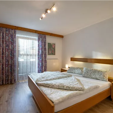 A cozy bedroom with a double bed and wooden furniture. Large windows with curtains provide plenty of daylight.