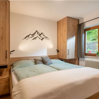 Ein gemütliches Schlafzimmer mit einem Doppelbett und Holzmöbeln. Durch das Fenster ist eine grüne Außenansicht sichtbar.