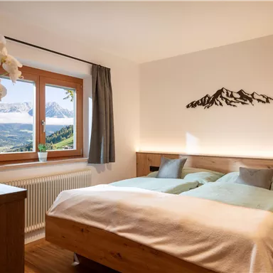 Ein modernes Schlafzimmer mit einem Doppelbett und sanften Farben. Im Hintergrund sind Fenster mit Blick auf die Berge und eine elegante Orchidee auf dem Nachttisch.