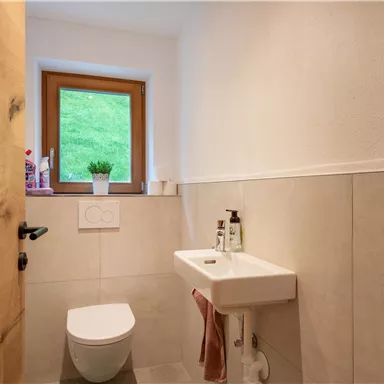 Ein modernes Badezimmer mit einem Waschbecken und einer Toilette. Es gibt ein Fenster, das Tageslicht und einen Blick ins Grüne bietet.