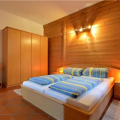 A cozy bedroom with a double bed and colorful blankets. The walls are made of wood and there is a wardrobe as well as a chair.