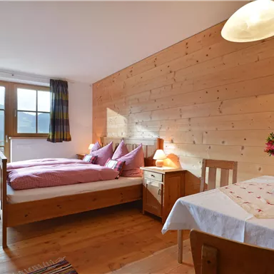 Ein gemütliches Schlafzimmer mit Holzdekor und einem großen Fenster. Der Raum ist hell beleuchtet und hat ein Bett mit lila Bettwäsche sowie einen kleinen Tisch mit Blumen.