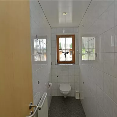 A bright bathroom with white tiles and a window. The sanitary facilities are simple and modern.