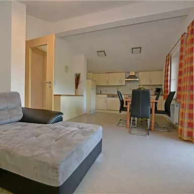 A cozy living room with a gray couch and a dining table. The kitchen is open and bright with large windows.