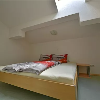 A simple bedroom with a double bed and two pillows. The room has a light wall and a small window opening for daylight.