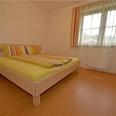 A bright bedroom with a double bed and colorful pillows. A large window with curtains lets in plenty of light.