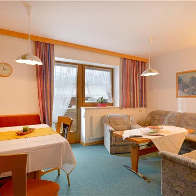 Ein gemütliches Wohnzimmer mit einer Sitzecke und einem Esstisch. Helle Farben und ein Fenster sorgen für eine einladende Atmosphäre.