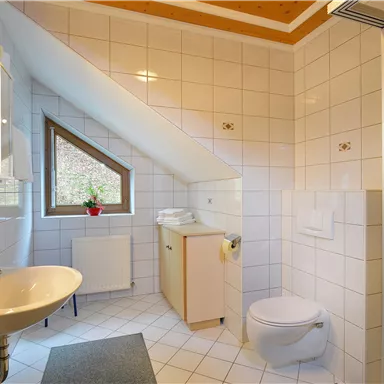 Ein modernes Badezimmer mit weißen Fliesen und einer schrägen Decke. Es verfügt über ein Waschbecken, eine Toilette und ein Fenster mit Blick nach draußen.