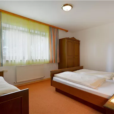 Ein schlichtes Schlafzimmer mit zwei Betten und einem Schrank. Die Fenster sind mit hellen Vorhängen dekoriert und der Boden ist mit Teppich ausgelegt.