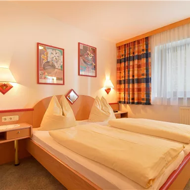 Ein gemütliches Schlafzimmer mit zwei Betten und bunten Kissen. Die Wände sind mit Kunstwerken dekoriert und das Fenster lässt viel Tageslicht herein.