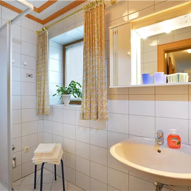 Ein modernes Badezimmer mit einer Duschkabine und einem Waschbecken. Es hat helle Fliesen und eine kleine Pflanze am Fenster.