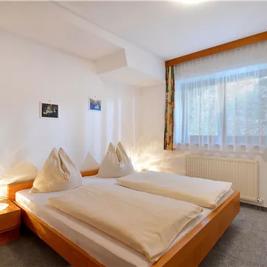 Ein helles Schlafzimmer mit zwei Betten und gemütlicher Bettwäsche. Der Raum hat große Fenster mit Vorhängen und eine warme Atmosphäre.