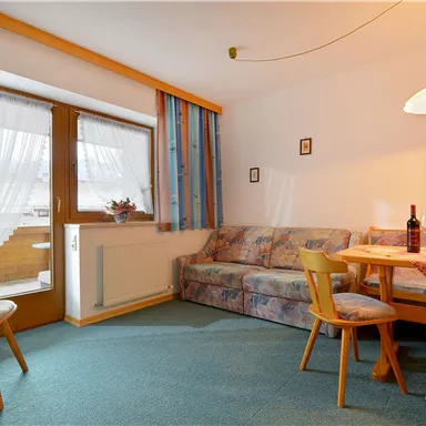 Ein gemütlicher Wohnraum mit einem Sofa, einem Tisch und Stühlen. Große Fenster sorgen für Tageslicht und eine einladende Atmosphäre.