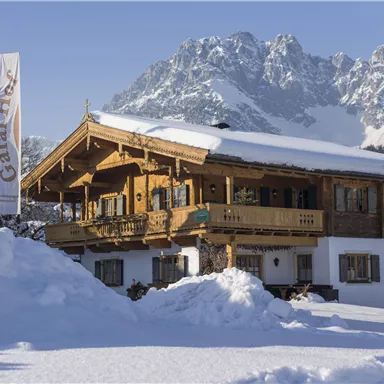 Ein traditionelles Holzhaus im Schnee, umgeben von Bergen. Die Szene zeigt eine ruhige Winterlandschaft.