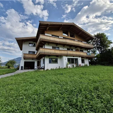 Ein modernes Haus mit mehreren Balkonen und einer großen Wiese davor. Der Himmel ist teilweise bewölkt und es gibt viel grünes Gras.