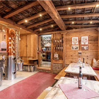 A cozy room with rustic decor and wooden walls. A distillery stands in the foreground, while tables with red tablecloths invite seating.