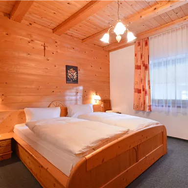 A cozy bedroom with wooden walls and a double bed. The room has large windows with curtains and warm light.