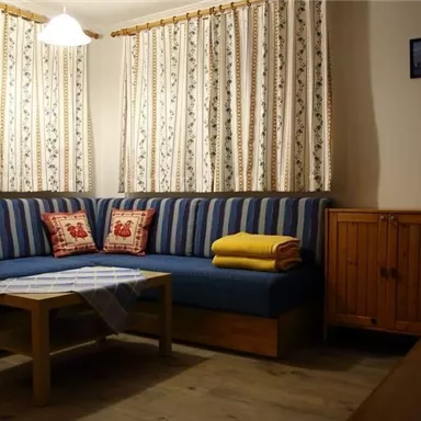 Ein gemütlicher Wohnraum mit einer blauen Couch und Kissen. Der Tisch ist mit einer Tischdecke gedeckt, und im Hintergrund sind Vorhänge mit Blumenmuster.