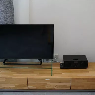 A modern television stands on a wooden TV rack with a glass top. To the right of the television is a black sound system.