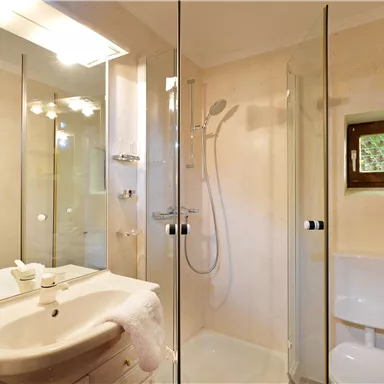 A modern bathroom with a glass shower and a white sink. The walls are in warm, soft tones and there is a small window.