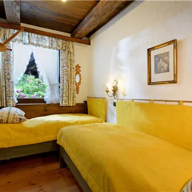 A cozy bedroom with two yellow beds and a wooden clothes rack. The window offers a view of a green garden.