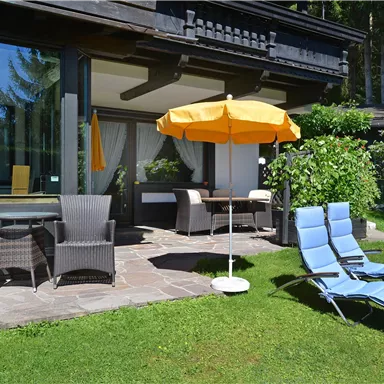 A cozy terrace with a dining table, seating, and an umbrella. In the foreground, there are deck chairs on the green lawn.