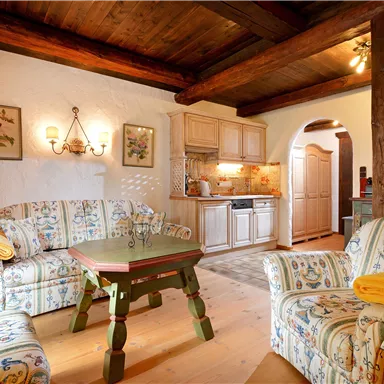 A cozy living space with a wooden beam ceiling and two comfortable sofas. The area is bright and inviting, with a small dining table and a fully equipped kitchen in the background.