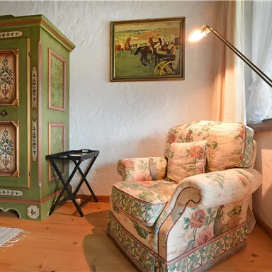 A cozy room with a floral patterned armchair and a green cabinet. A picture hangs on the wall, and a floor lamp provides additional light.