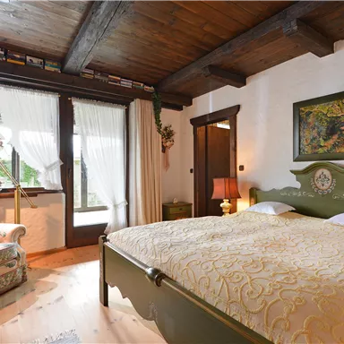 A cozy bedroom with a large bed and a chair in the corner. The windows are bright and offer a view outside.