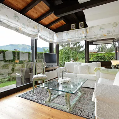 A bright and welcoming living room with large windows overlooking the garden. It features a cozy sofa area and a glass table.