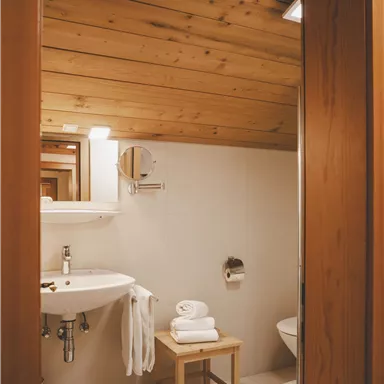 Ein modernes Badezimmer mit Holzdecke und hellen Fliesen. Es gibt ein Waschbecken, Handtücher und einen Hocker.