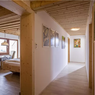 A bright hallway with wooden ceilings and walls. Pictures hang on the walls, and at the end of the hallway, a bedroom is visible.