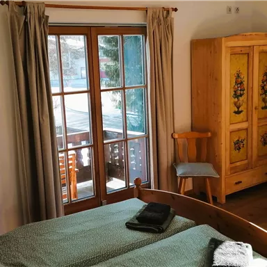 Ein gemütliches Schlafzimmer mit einem Bett, einem Sofa und einem schönen Holzkasten. Große Fenster bieten viel Tageslicht und Ausblick auf den Balkon.