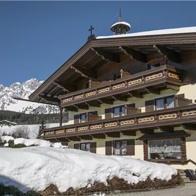 Ein traditionelles alpines Haus im Winter mit Schnee bedeckt. Im Hintergrund sieht man majestätische Berge unter klarem Himmel.