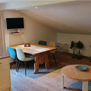 Ein helles und einladendes Wohnzimmer mit einer gemütlichen Sitzecke. Am Tisch stehen Stühle und der Raum hat große Fenster, die viel Tageslicht hereinlassen.