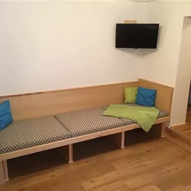 Ein schlichtes Wohnzimmer mit einer Bänke und bunten Kissen. An der Wand hängt ein Fernseher, und der Boden ist aus Holz.