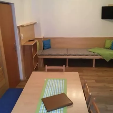 Ein einfaches Esszimmer mit einem Holztisch und Stühlen. An der Wand befindet sich eine Sitzecke und ein Fernsehgerät.