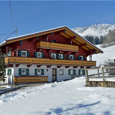 A beautiful red and white painted farmhouse in the snow. In the background, there are snow-covered mountains and a clear blue sky.