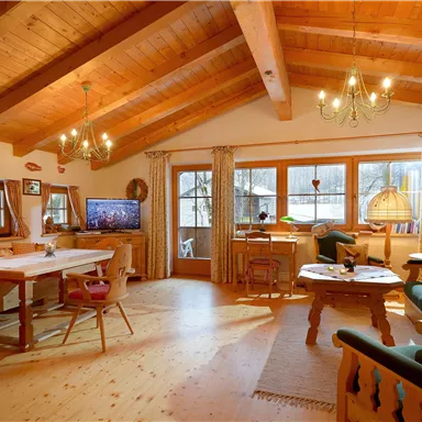 A cozy living room with wooden ceilings and large windows. The room is furnished with a dining table, comfortable sofas, and a TV corner.