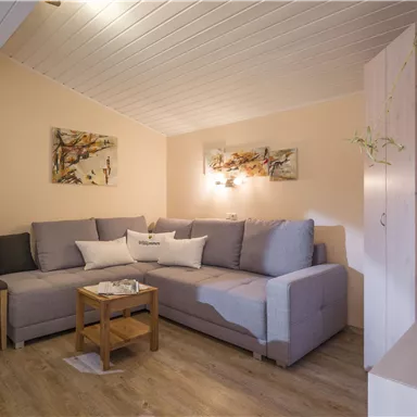 A cozy living room with a gray couch and a small wooden table. Pictures hang on the walls, and there is a pleasant, warm lighting.