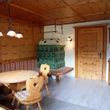 Ein gemütlicher Raum mit Holzverkleidung und einer Sitzecke. Im Hintergrund steht ein traditioneller Kochofen und eine Holztür.