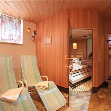 Ein gemütlicher Wellnessbereich mit Liegen und einer Sauna im Hintergrund. Die Wände sind aus Holz, und es gibt helle Fenster mit Vorhängen.