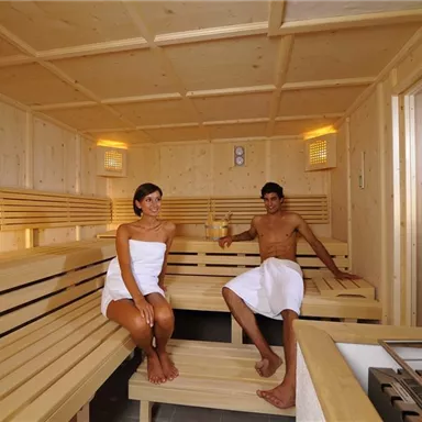Ein modernes Saunainneres mit Holzvertäfelung. Zwei Personen entspannen in Handtüchern auf den Bänken.