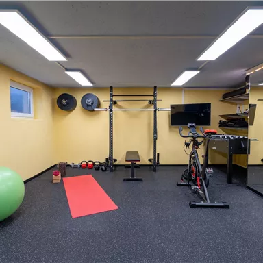 Ein moderner Fitnessraum mit gelben Wänden und einer schwarzen Gummibodenfläche. Es sind verschiedene Trainingsgeräte, eine Trainingsmatte und ein Fitnessball vorhanden.