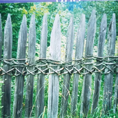 A rustic wooden fence with pointed posts and woven material in the middle. Surrounded by green grass and plants.