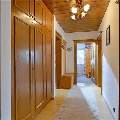 Ein heller Flur mit Holzvertäfelungen und einem warmen Beleuchtungskörper. Am Ende des Flurs sind Türen und ein Teppich zu sehen.