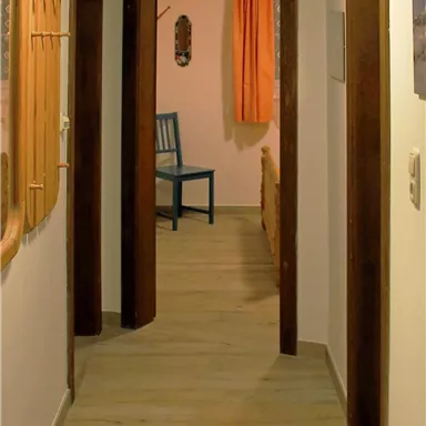 Ein heller Flur mit mehreren Türen und einem blauen Stuhl am Ende. Die Wände sind in neutralen Farben gehalten und es gibt orange Vorhänge.