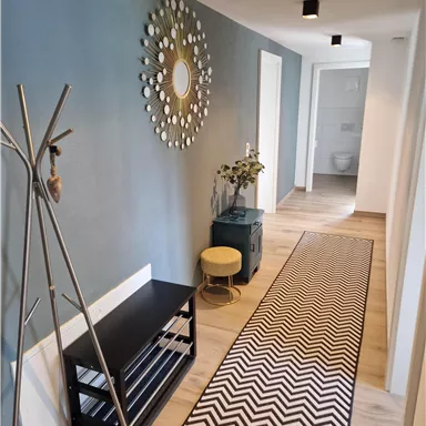 A modern hallway with a gray wall and a stylish mirror. A rug with a chevron pattern lies on the floor, and there are some pieces of furniture, including a wardrobe and a small table.