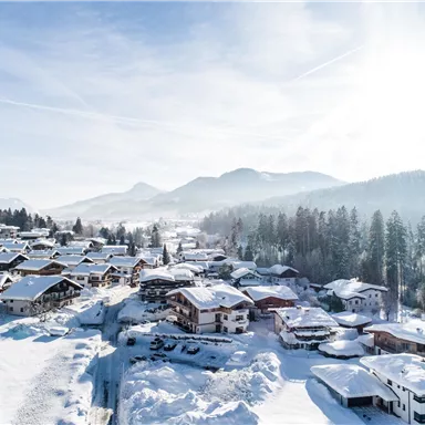 A picturesque winter landscape with snow-covered houses and trees. The sun shines over the mountains in the background.