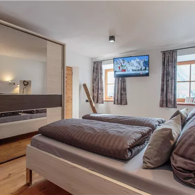 Ein modernes Schlafzimmer mit einem komfortablen Bett und einem Spiegelkleiderschrank. Ein Fernseher hängt an der Wand und es gibt Fenster mit Vorhängen, die natürliches Licht hereinlassen.